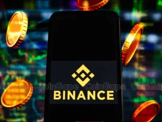 Smartphone mostrando o logotipo dourado da Binance sobre fundo preto, cercado por moedas douradas em um ambiente com gráficos de mercado coloridos ao fundo.