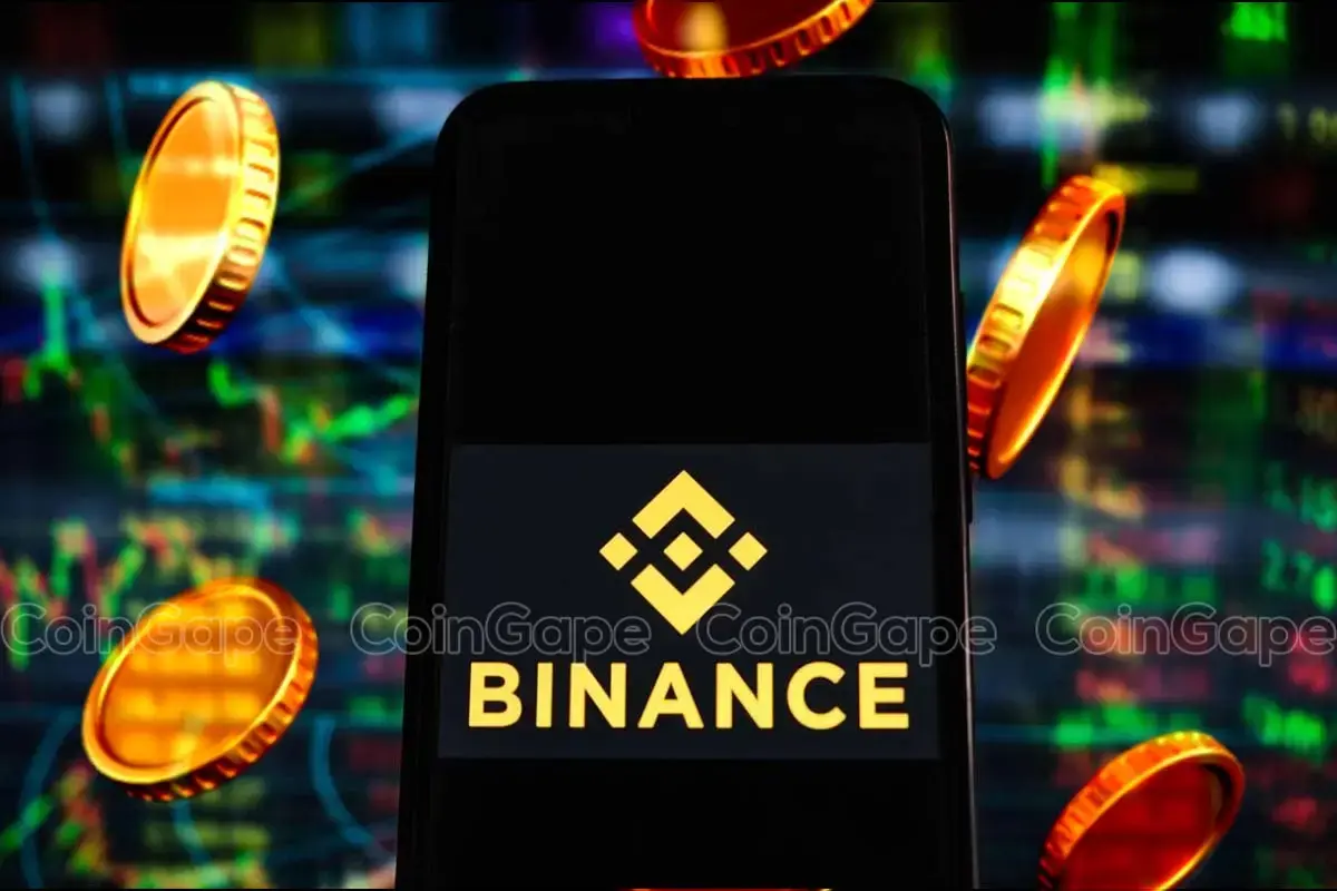 Smartphone mostrando o logotipo dourado da Binance sobre fundo preto, cercado por moedas douradas em um ambiente com gráficos de mercado coloridos ao fundo.