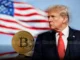 Presidente Donald Trump de perfil ao lado de uma moeda Bitcoin física com a bandeira dos Estados Unidos ao fundo.