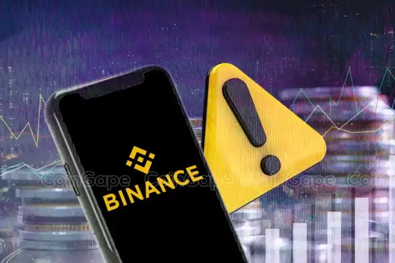 Smartphone exibindo logo da Binance ao lado de símbolo de alerta amarelo, com gráficos de mercado ao fundo.