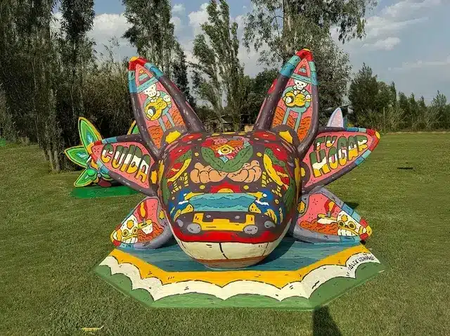Escultura colorida de axolotl em forma de flor com arte mexicana tradicional instalada em campo verde com árvores ao fundo.
