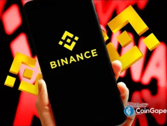 Mãos segurando smartphone com logo da Binance em fundo vermelho simbolizando queda no mercado cripto