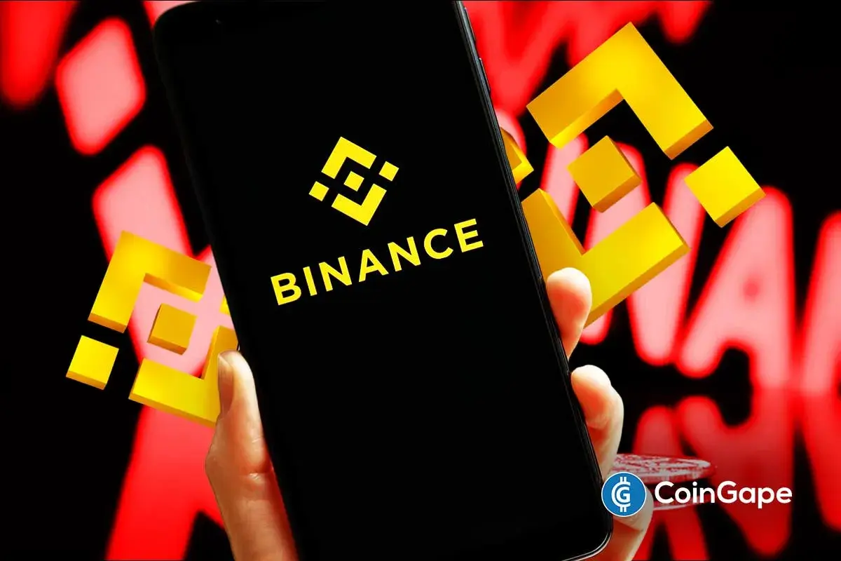 Mãos segurando smartphone com logo da Binance em fundo vermelho simbolizando queda no mercado cripto