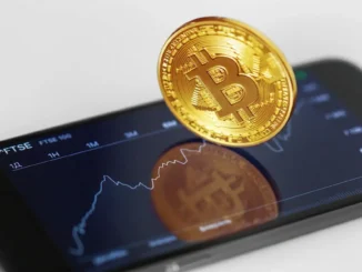 Moeda física de Bitcoin dourada posicionada sobre smartphone mostrando gráfico de preços de criptomoedas em exchanges brasileiras.