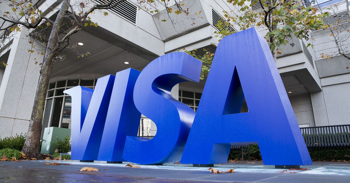 Logotipo da Visa em letras azuis tridimensionais na frente de um edifício corporativo.