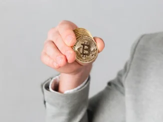 Mão vestida com paletó cinza segurando um conjunto de moedas douradas com o símbolo do bitcoin.